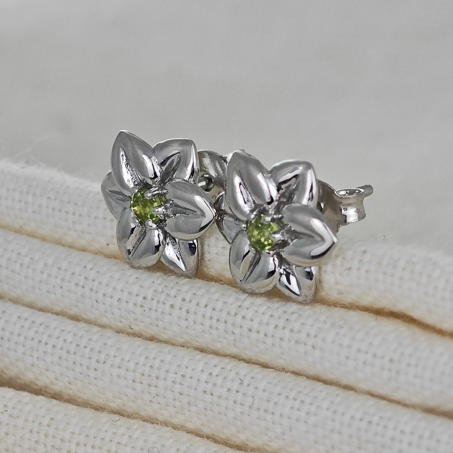 Silver flower-shaped earrings with green gemstones on a beige fabric background