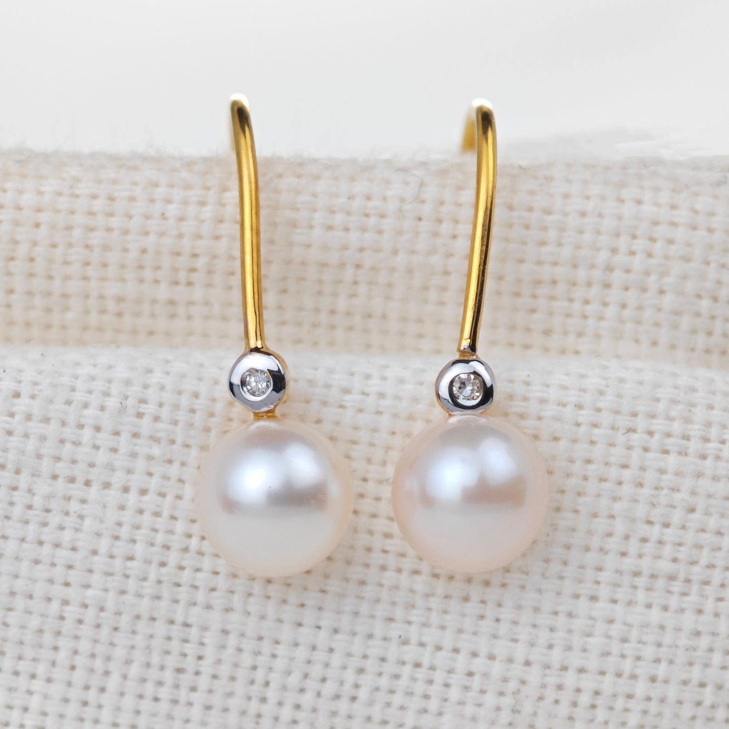 Pearl earrings with gold hooks and small diamonds on a beige fabric background