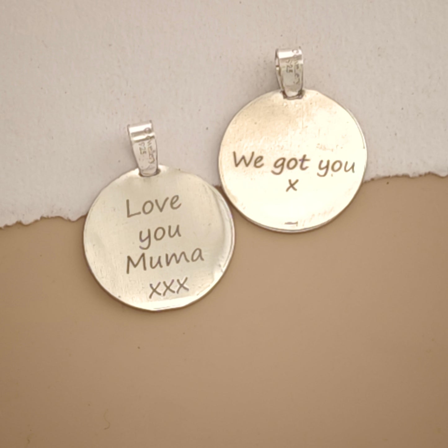 Two round silver pendant charms with engraved messages, one reading 'Love you Muma xxx' and the other 'We got you x', displayed on a split beige and white background.