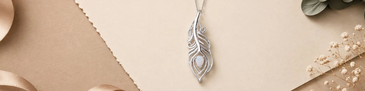 A silver-toned feather pendant necklace with a central white opal stone, displayed on a beige background with decorative ribbon and dried flowers.