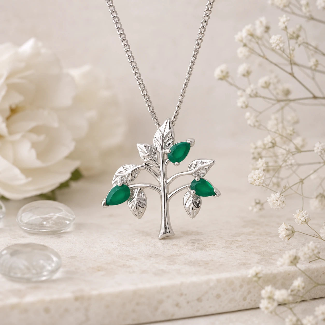 Silver tree-shaped pendant with green gemstones on a chain, set against a light background with flowers.
