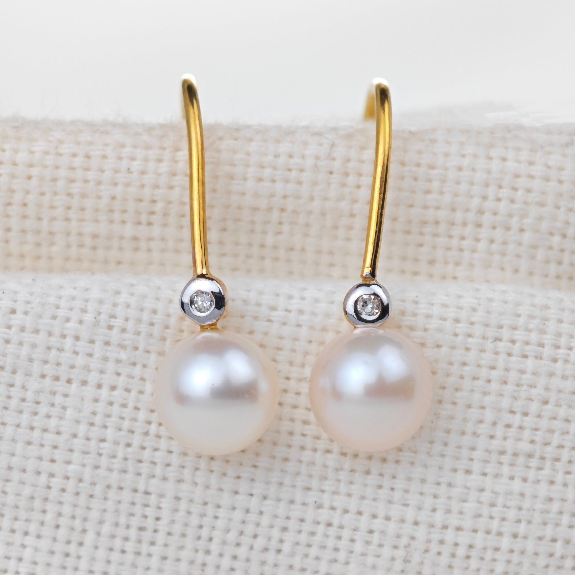 Pearl earrings with gold hooks and small diamonds on a beige fabric background