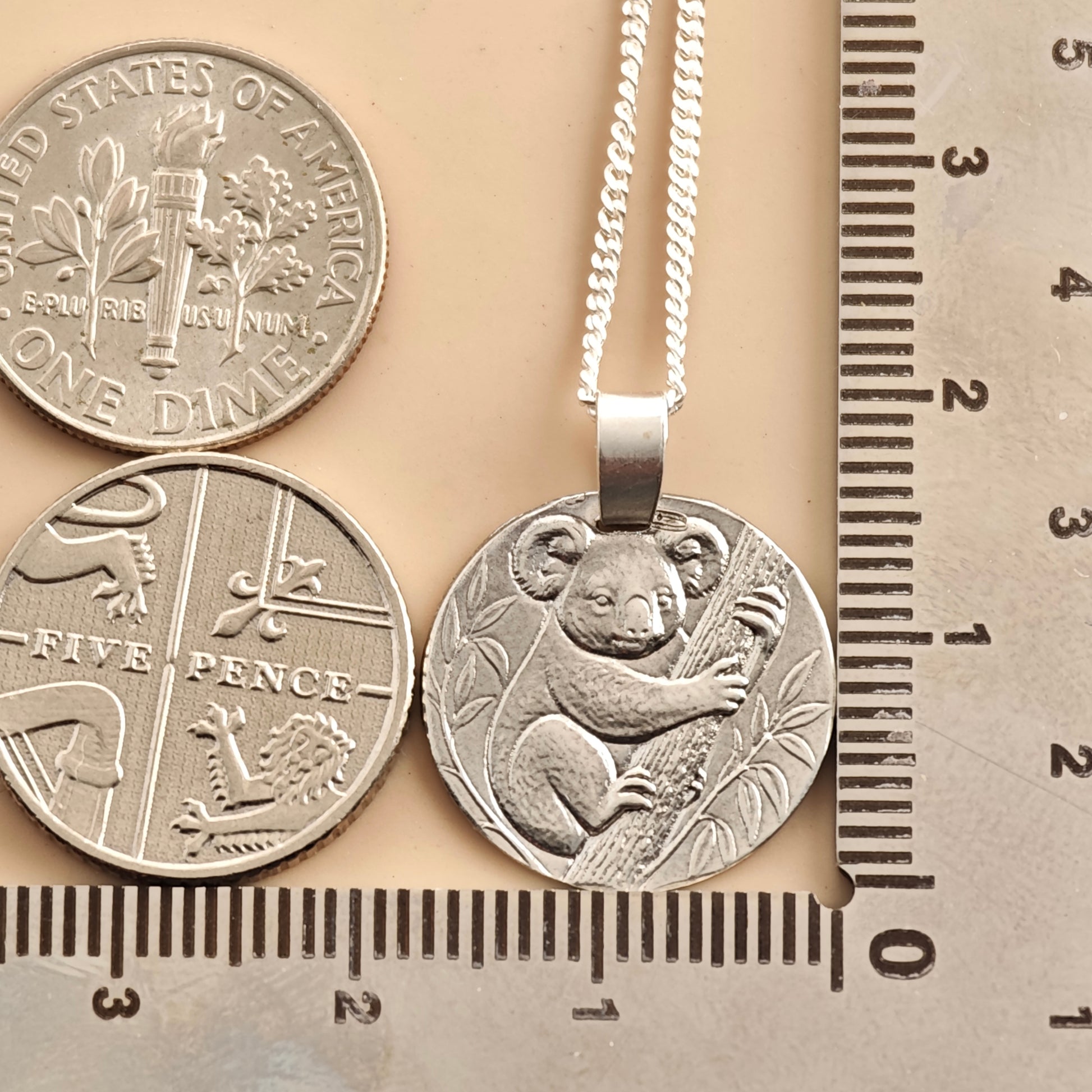 A silver pendant necklace featuring an embossed koala holding a eucalyptus branch, shown next to  a five pence piece and a US dime coin for scale on a beige background with a ruler on the right.