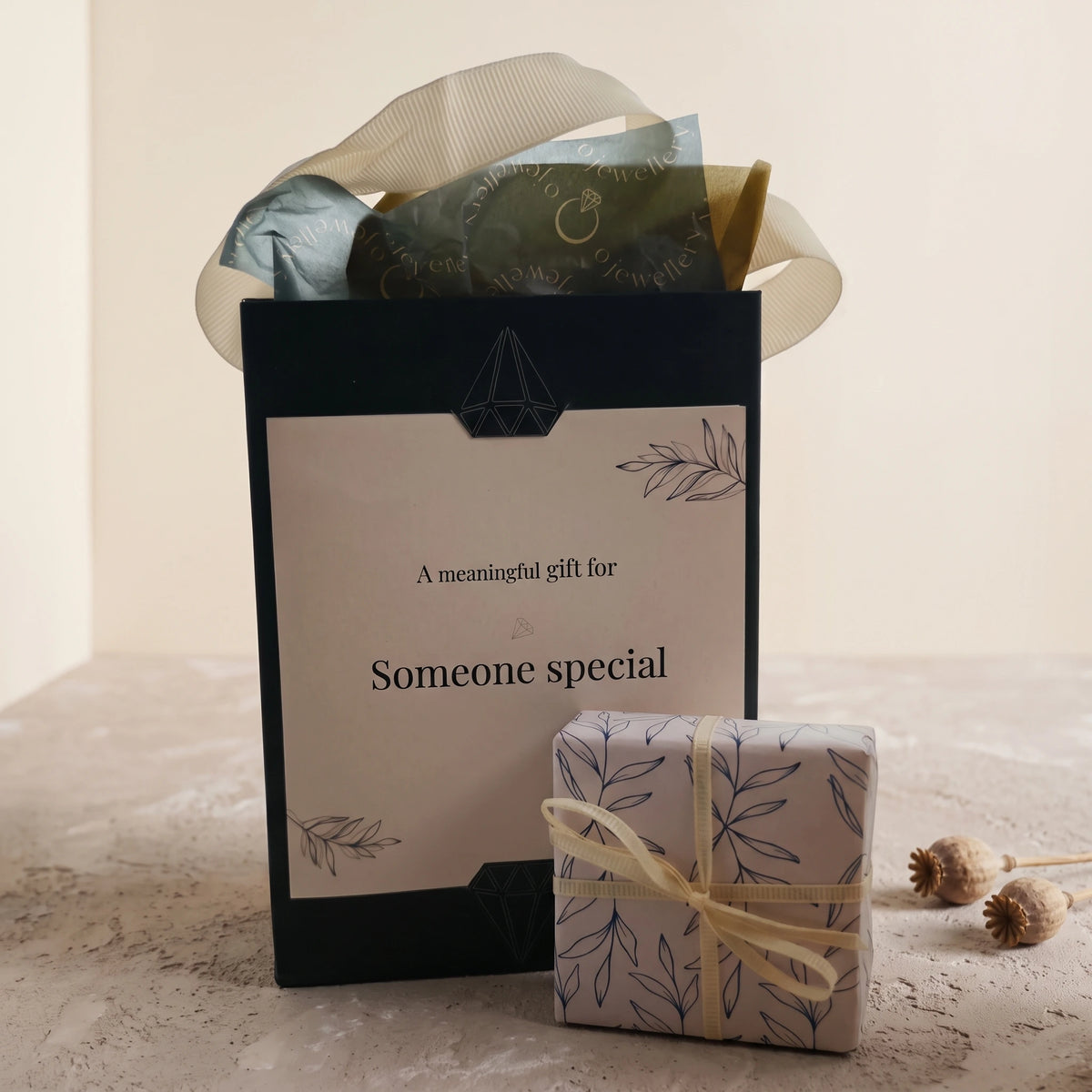 An Ojewellery gift set presentation featuring a tall forest green gift bag with a cream gift card insert that reads 'A meaningful gift for Someone special' and a small wrapped gift box with a matching leaf pattern and cream ribbon, styled on a neutral surface