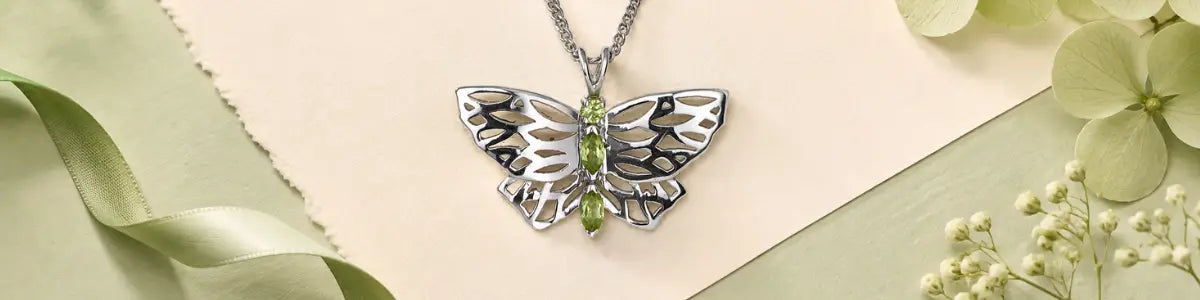 A silver butterfly pendant necklace with a green peridot gemstone center, displayed on a light beige background with decorative green ribbon and white flowers.