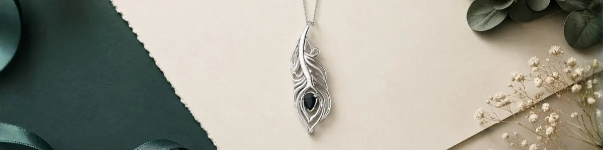A silver feather pendant with a dark blue stone in the center, hanging from a delicate chain. The pendant is styled in a flat-lay composition on a beige background with a dark green textured paper accent on the left and dried white flowers on the right.